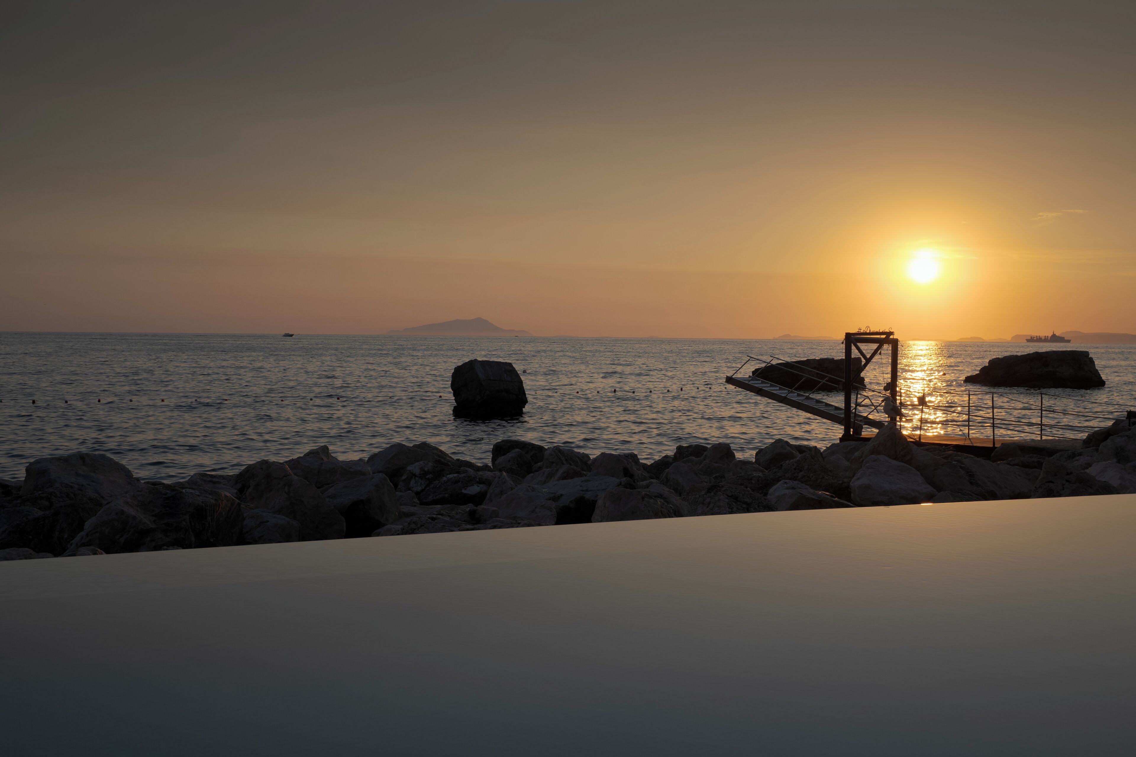 A sunset over a calm sea with rocky outcrops and a bridge structure.