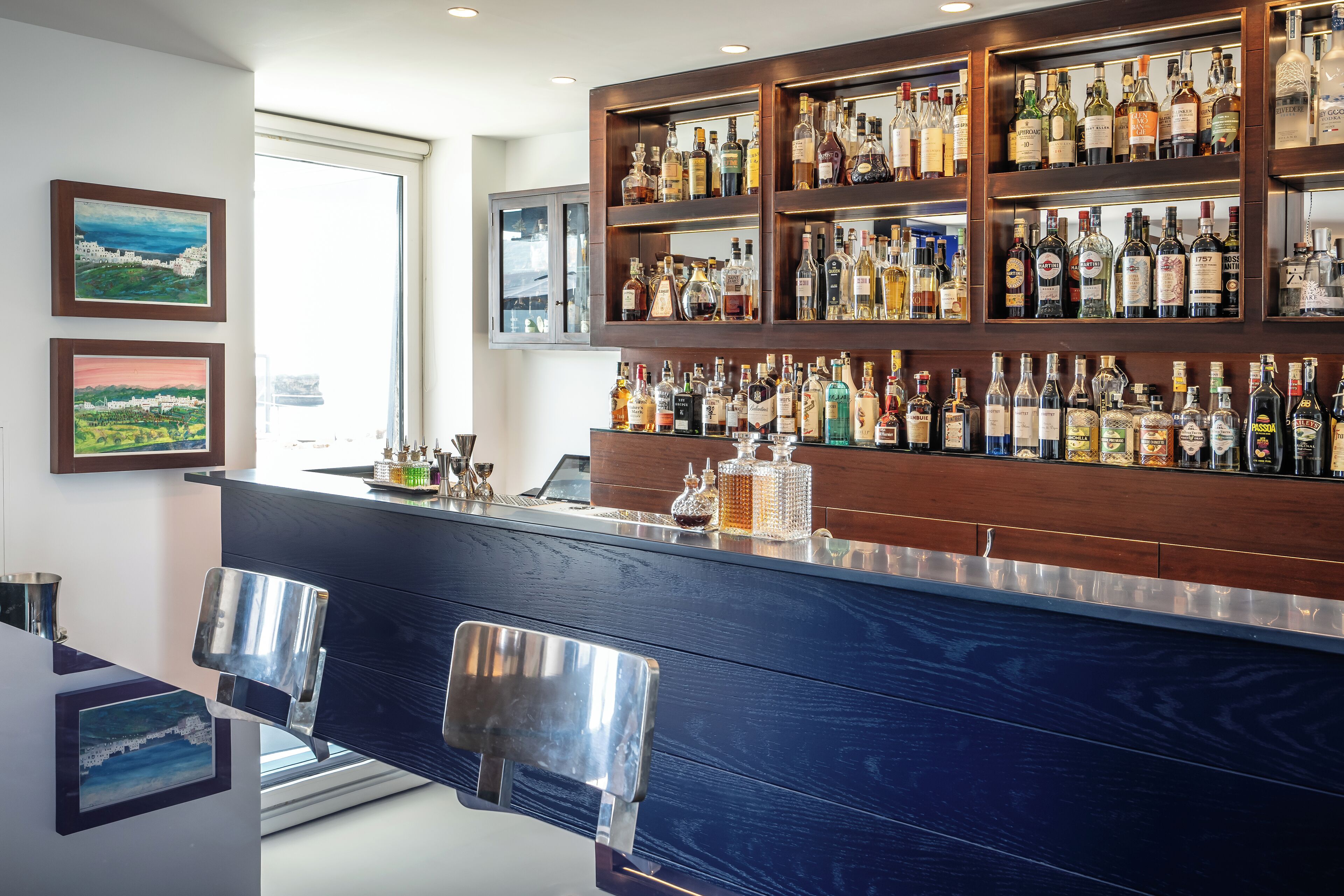Un espace bar avec une étagère en bois où sont rangées différentes bouteilles d’alcool, un comptoir bleu et deux chaises en métal.