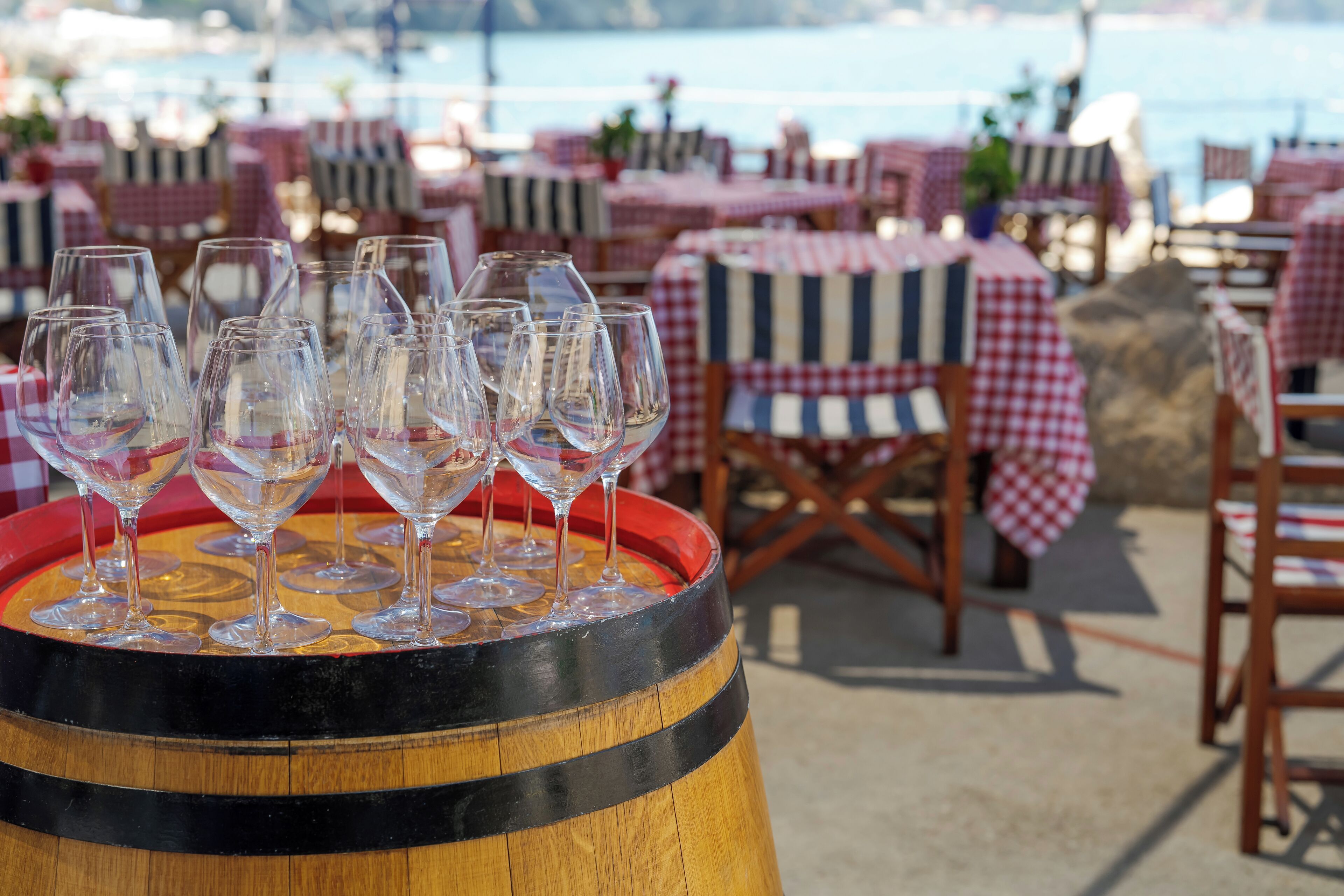 A wooden barrel with wine glasses arranged on top, surrounded by tables with checkered tablecloths and chairs, set against a coastal backdrop.