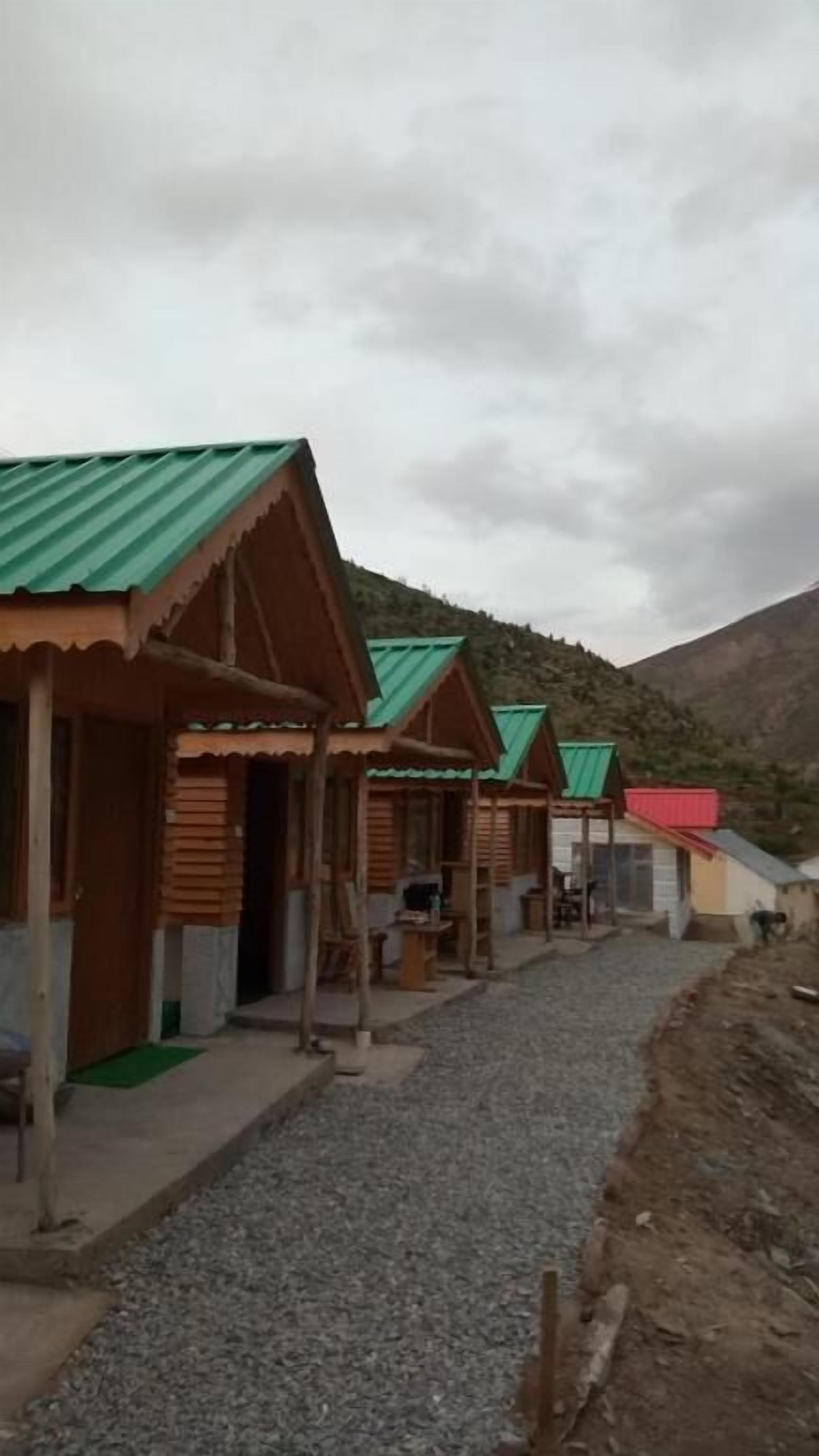 A row of wooden cabins with green roofs, arranged along a gravel path with a mountainous backdrop.
