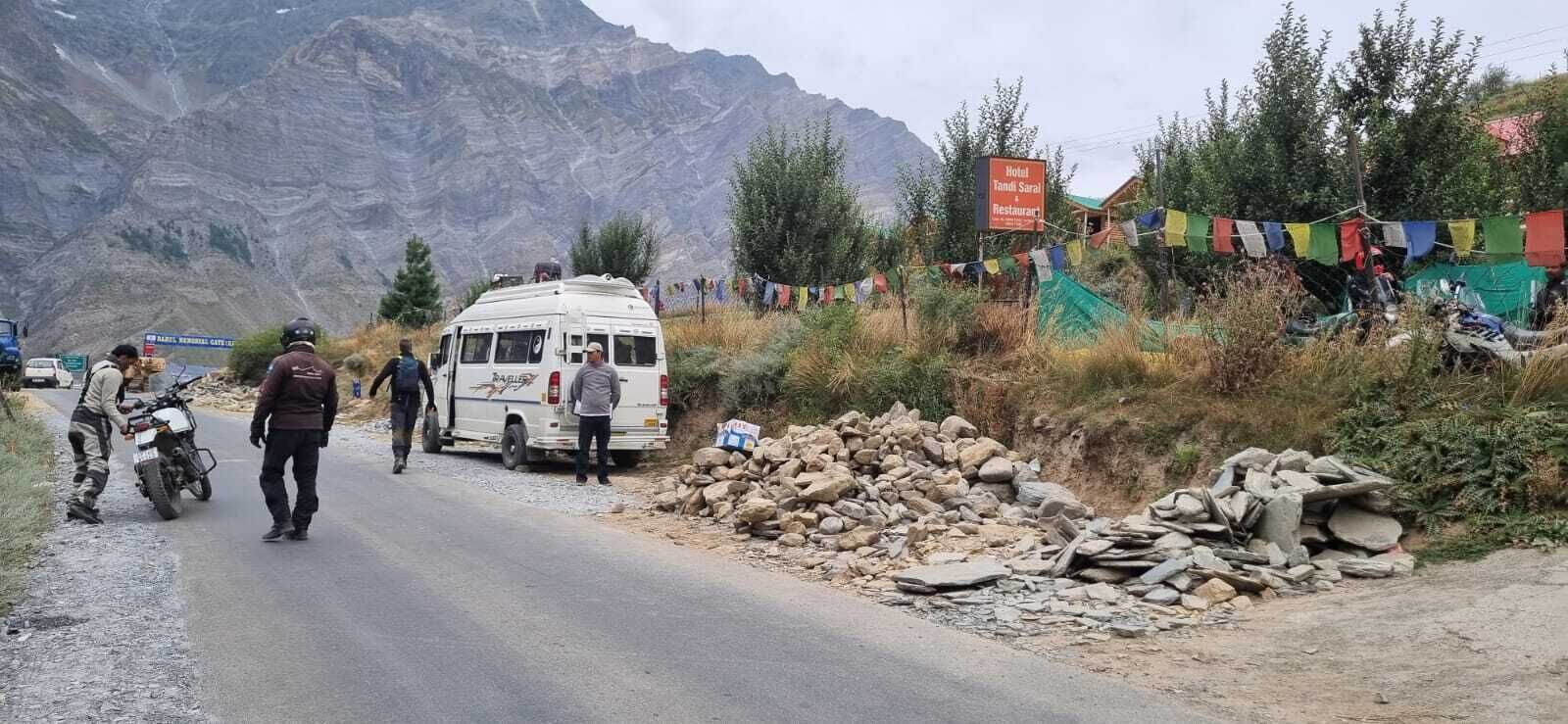 A road with a white van and motorcycles, a sign for Hotel Tandi Sarai Restaurant, and a mountainous backdrop.