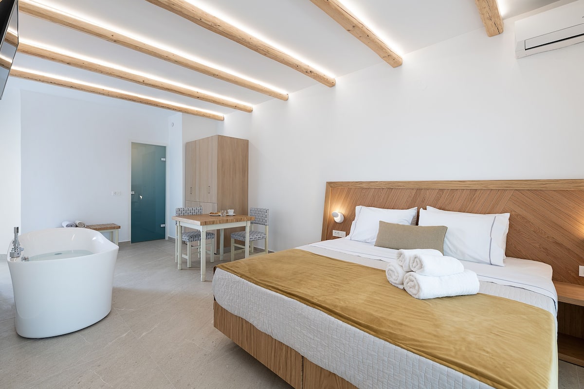 A modern hotel room with a large bed, a wooden headboard, a built-in bathtub, and a small desk with chairs.