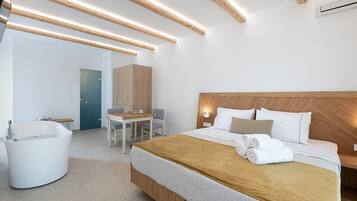 A modern hotel room with a large bed, a wooden headboard, a built-in bathtub, and a small desk with chairs.