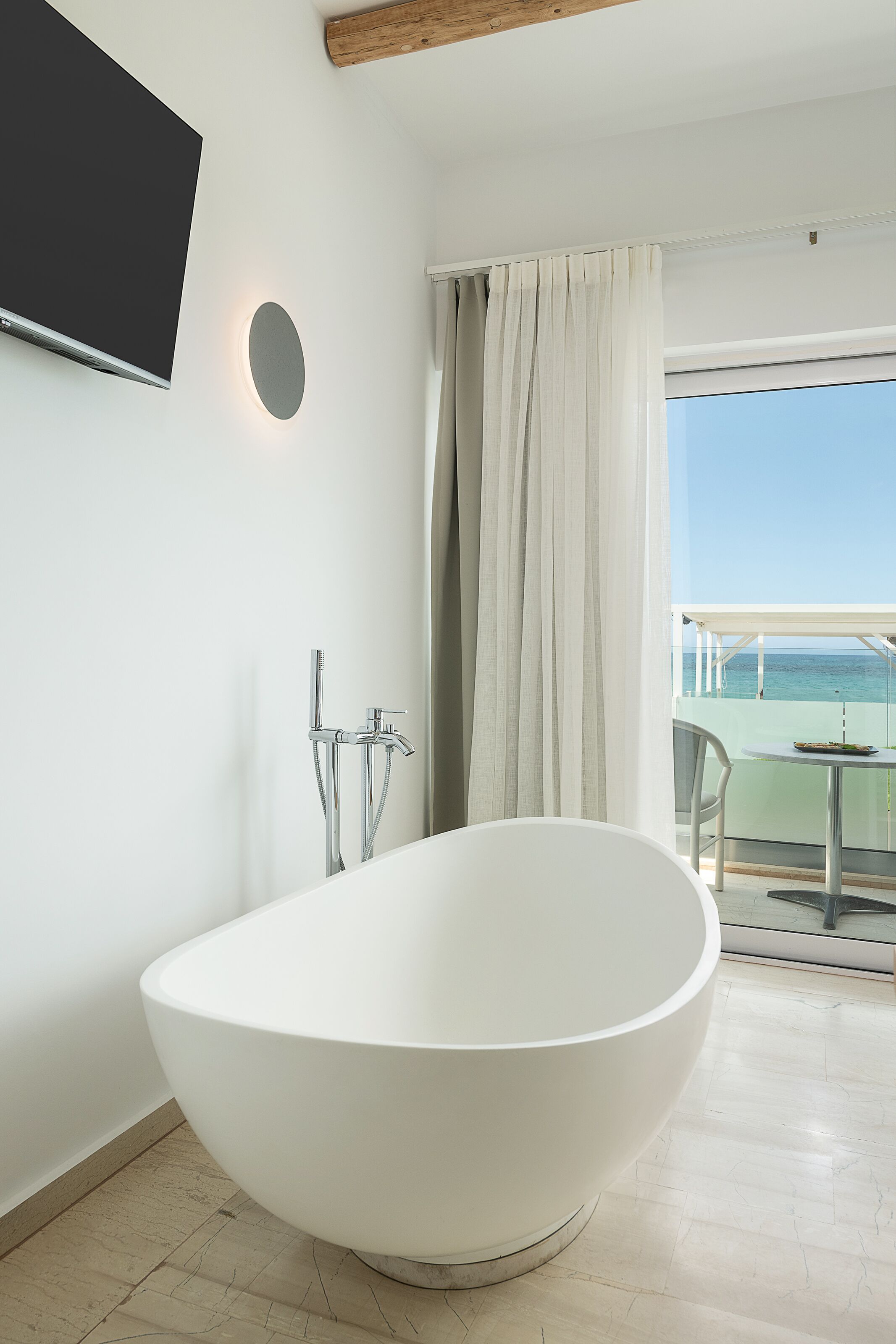 Een moderne badkamer met een vrijstaand bad, een wandgemonteerde televisie en uitzicht op zee door een groot raam.