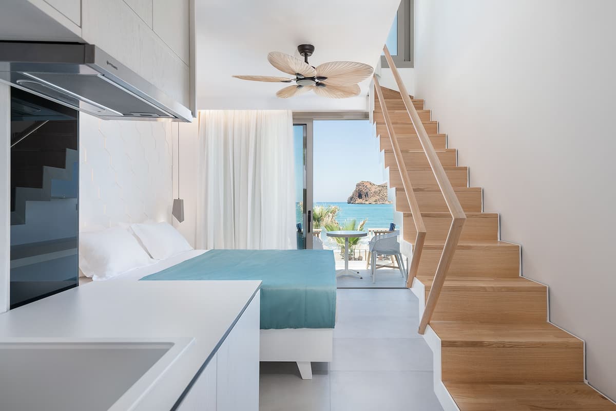 A modern hotel room with a bed, a desk, a ceiling fan, a staircase, and a view of the sea.