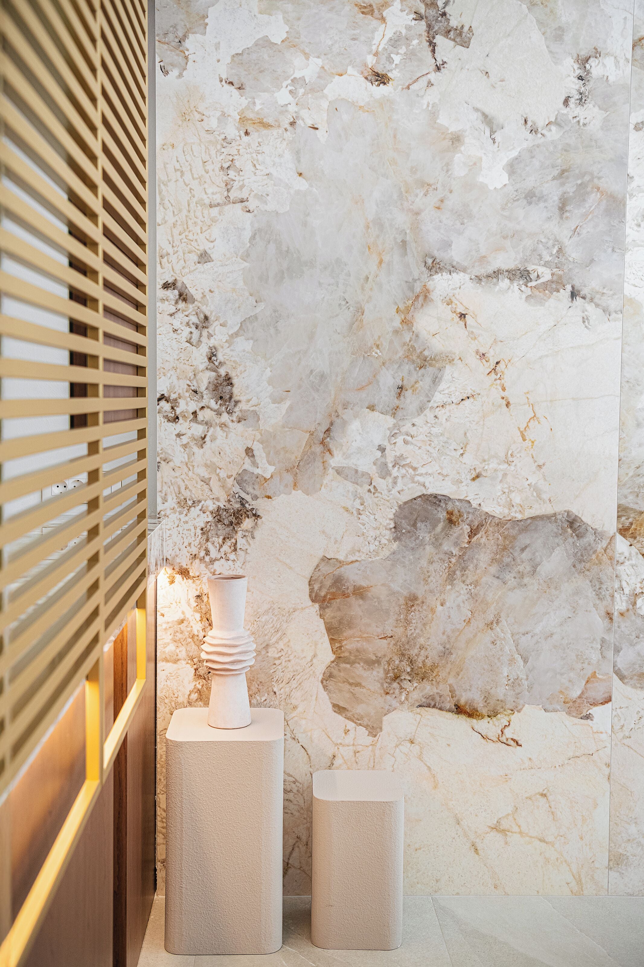 Un intérieur moderne avec un mur en marbre texturé, une cloison en lattes de bois et deux objets cylindriques blancs posés sur un sol carrelé.