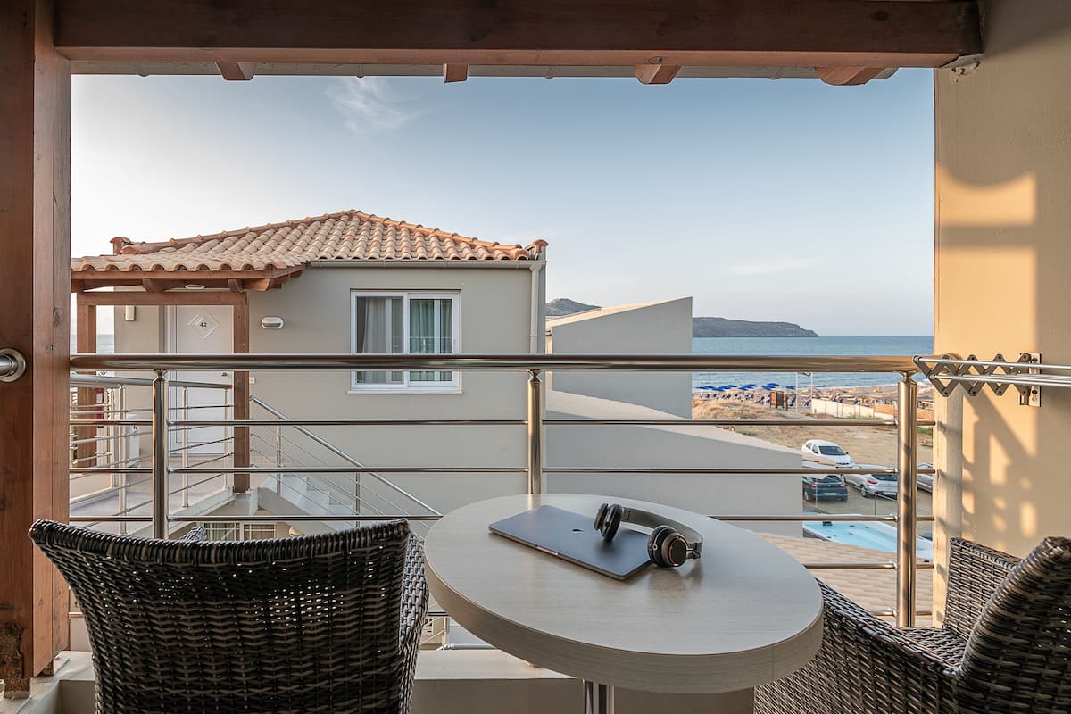 A balcony with wicker chairs, a table with a laptop and headphones, and a view of the beach and sea.
