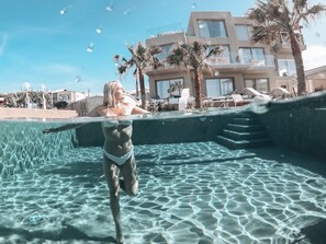 Una piscina con agua cristalina, una persona en traje de baño y un edificio moderno con balcones al fondo.