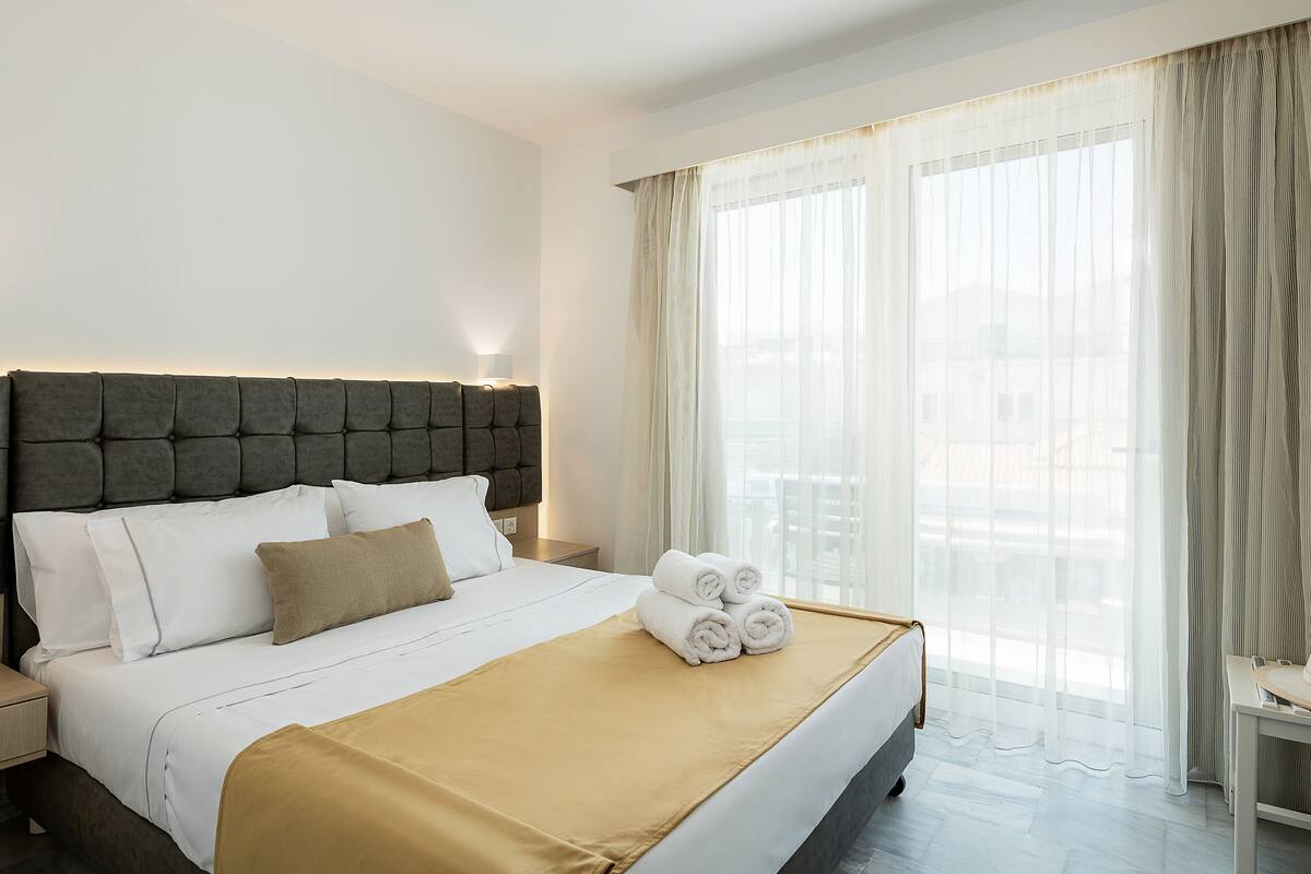 A hotel room with a large bed, a headboard, a bedside table, and a view of the city through sheer curtains.