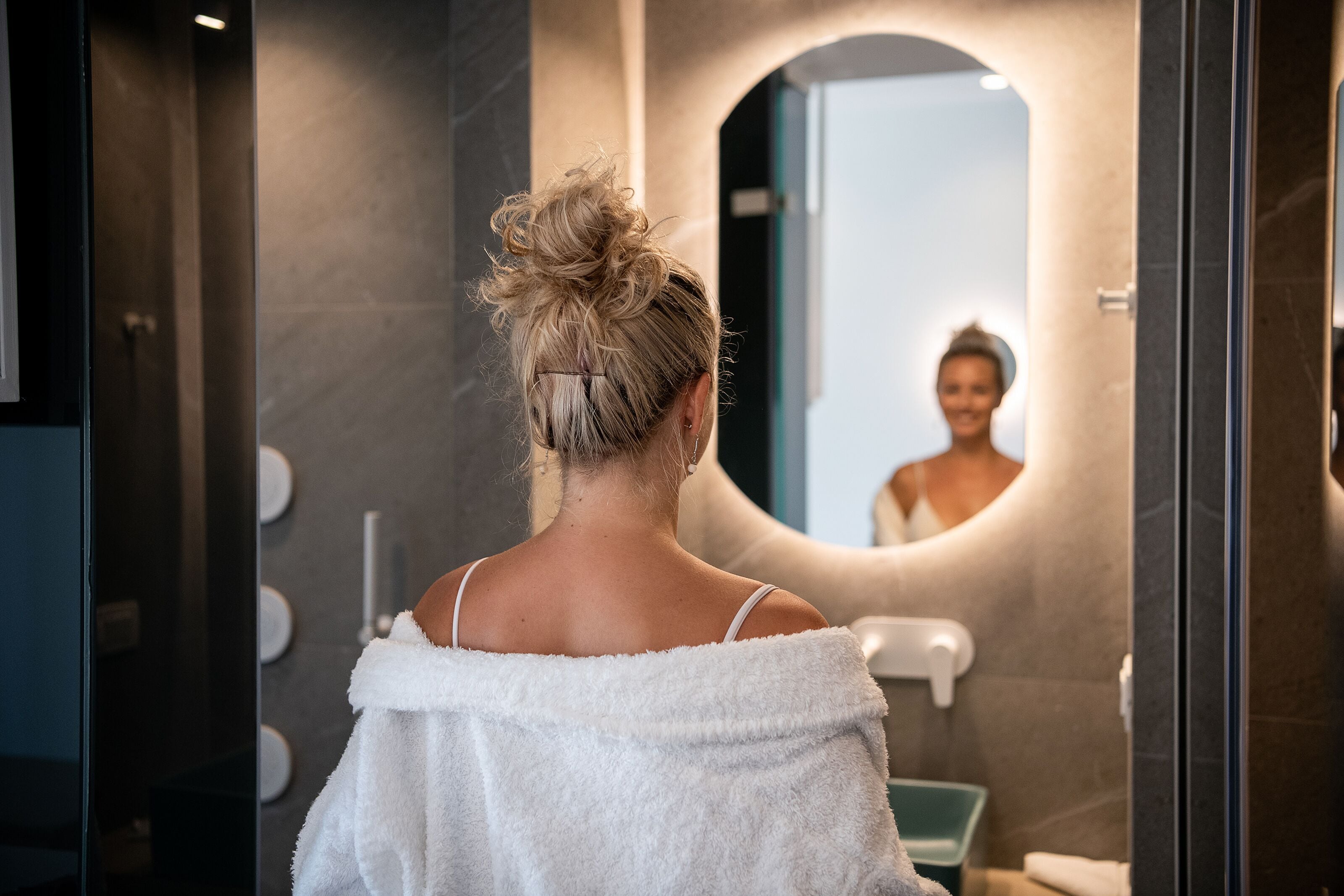 A person in a bathroom with a large mirror, a towel draped over their shoulders, and a sink with a faucet.