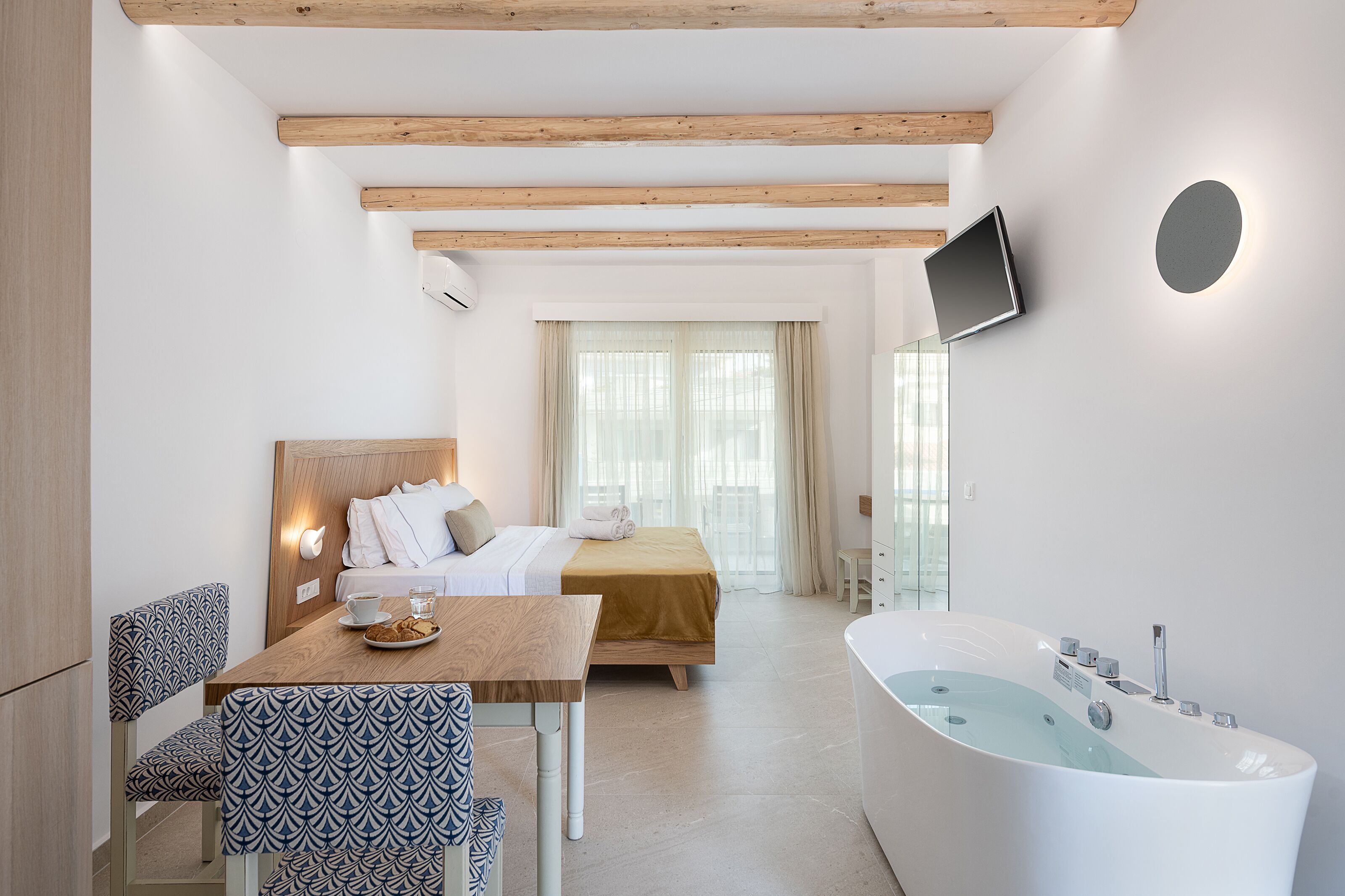 Une chambre d’hôtel moderne avec un plafond en bois, un lit recouvert d’une courtepointe beige, un bureau blanc sur lequel se trouve une assiette de biscuits, une grande baignoire et un téléviseur fixé au mur.