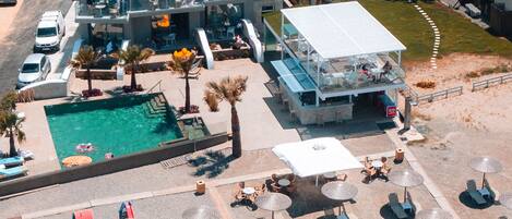 An aerial view of a hotel with a pool, multiple dining areas, and a beach with sun loungers and umbrellas.