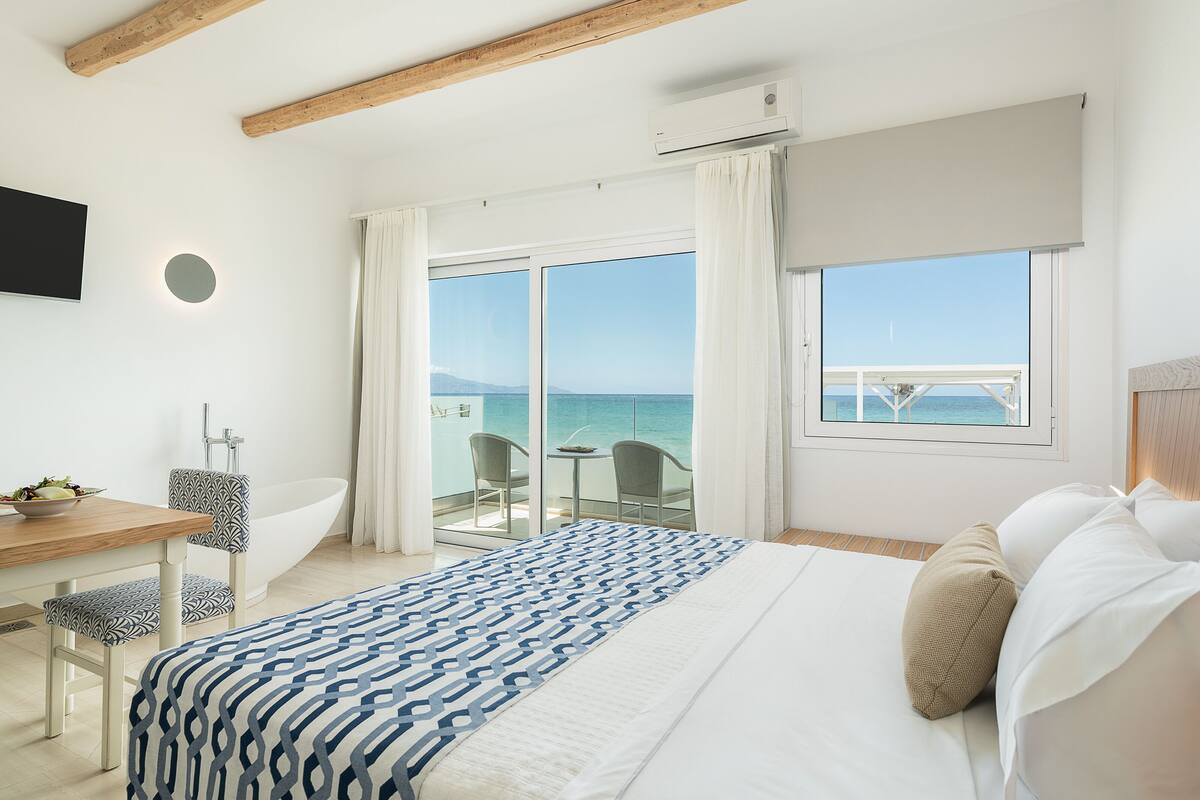 A bedroom with a large bed, a desk with a chair, a bathtub, and a balcony with a view of the sea.