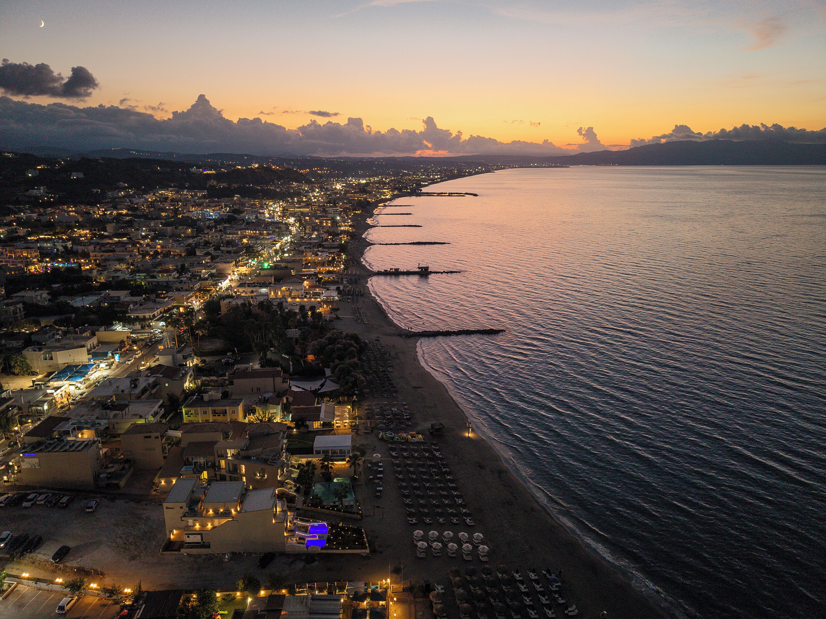 An aerial view of a coastal town at sunset with a beach, boats, and a marina.
