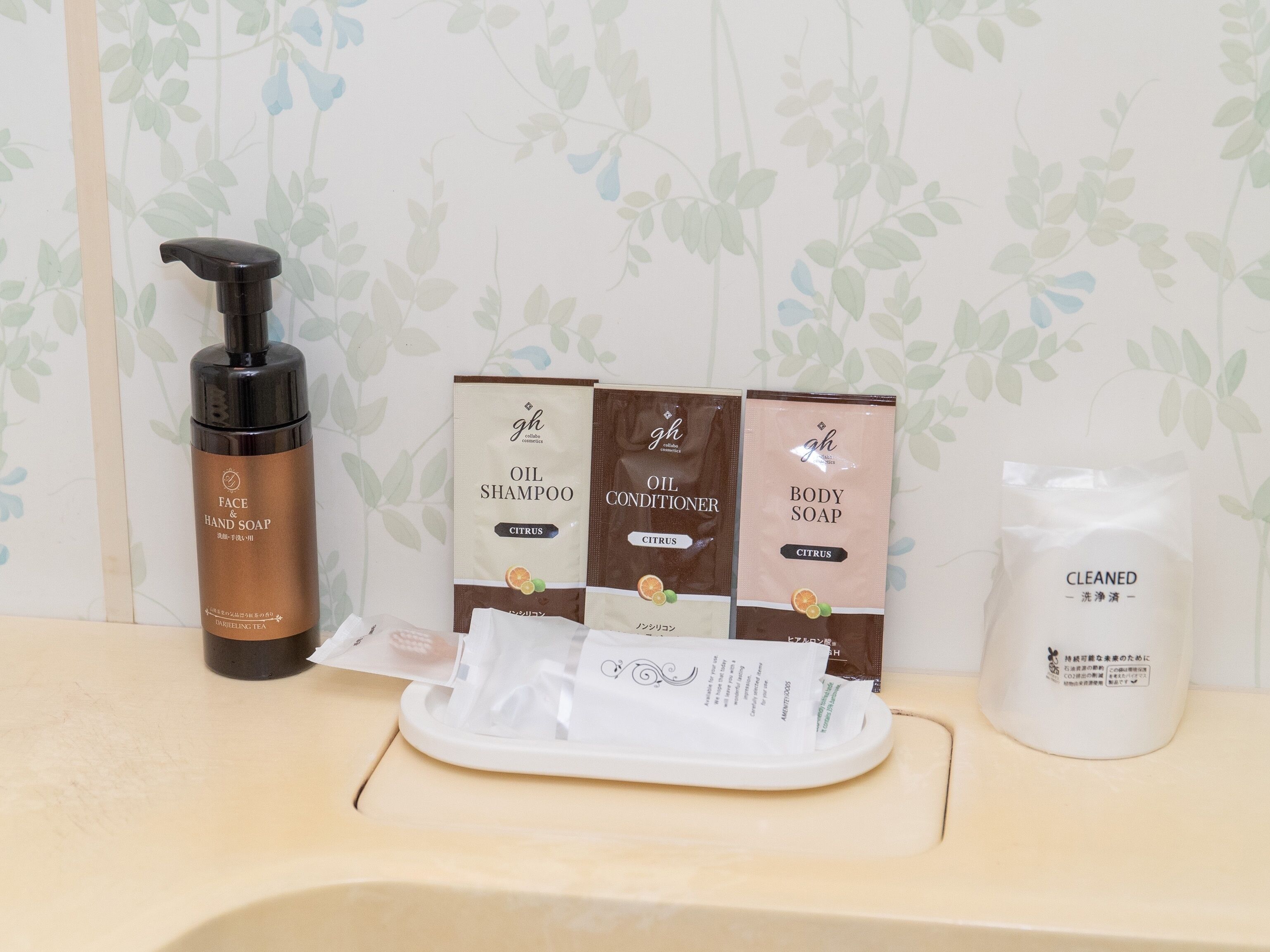 A bathroom counter with a bottle of face and hand soap, a bottle of oil shampoo, a body soap, and a container of cleaned water.
