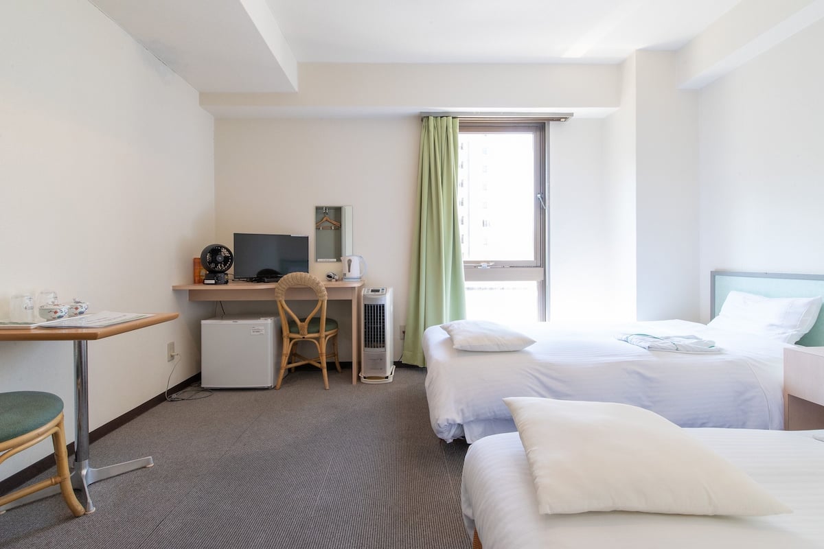 Une chambre d’hôtel équipée d’un bureau, de deux lits, d’une chaise, d’une petite table et d’une fenêtre avec des rideaux.