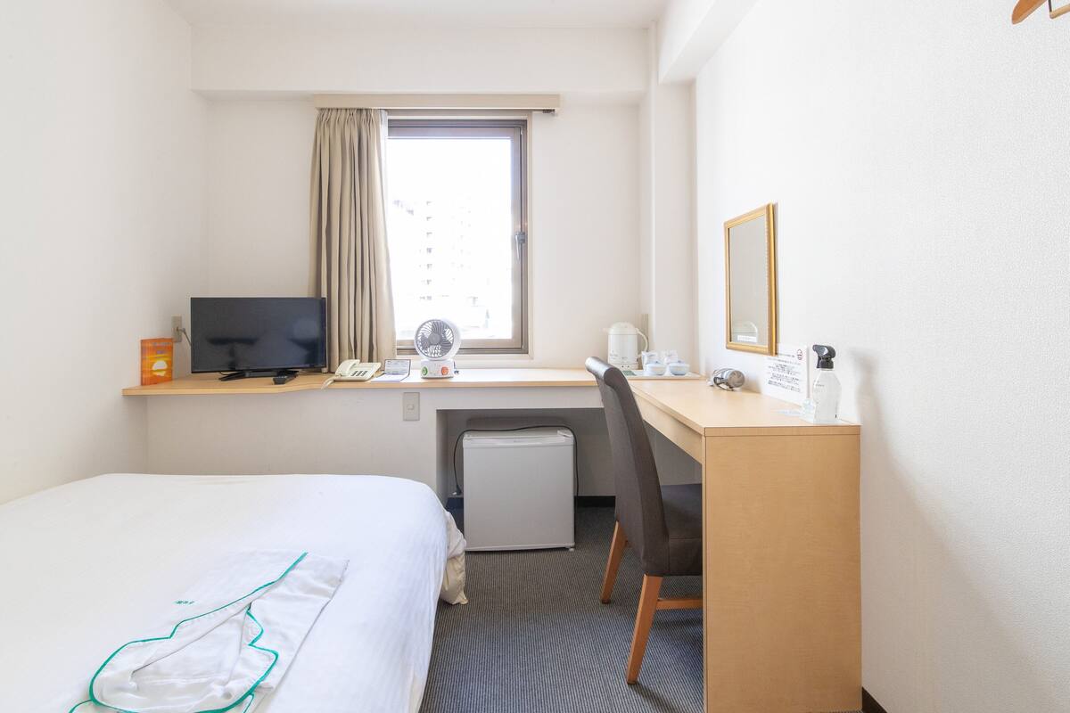 Une chambre d’hôtel comprenant un lit, un bureau avec un ordinateur, une chaise, un petit réfrigérateur, une fenêtre avec des rideaux et un miroir.