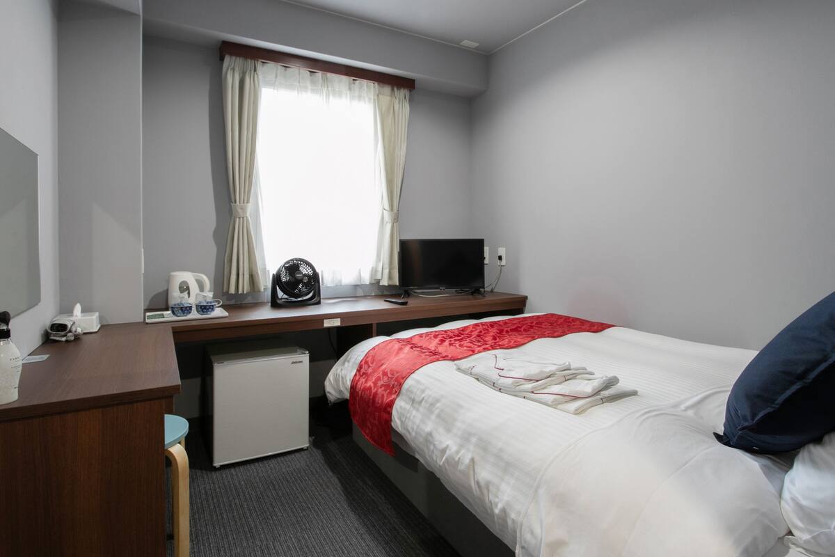 Une chambre d’hôtel comprenant un lit, un bureau avec un ordinateur, un petit réfrigérateur, une fenêtre avec des rideaux et une table de chevet avec une bouilloire et des tasses.