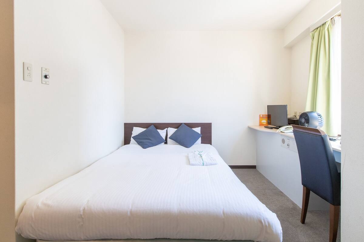Une chambre d’hôtel comprenant un lit, un bureau, une chaise, une lampe de bureau, un ventilateur et une fenêtre avec des rideaux.