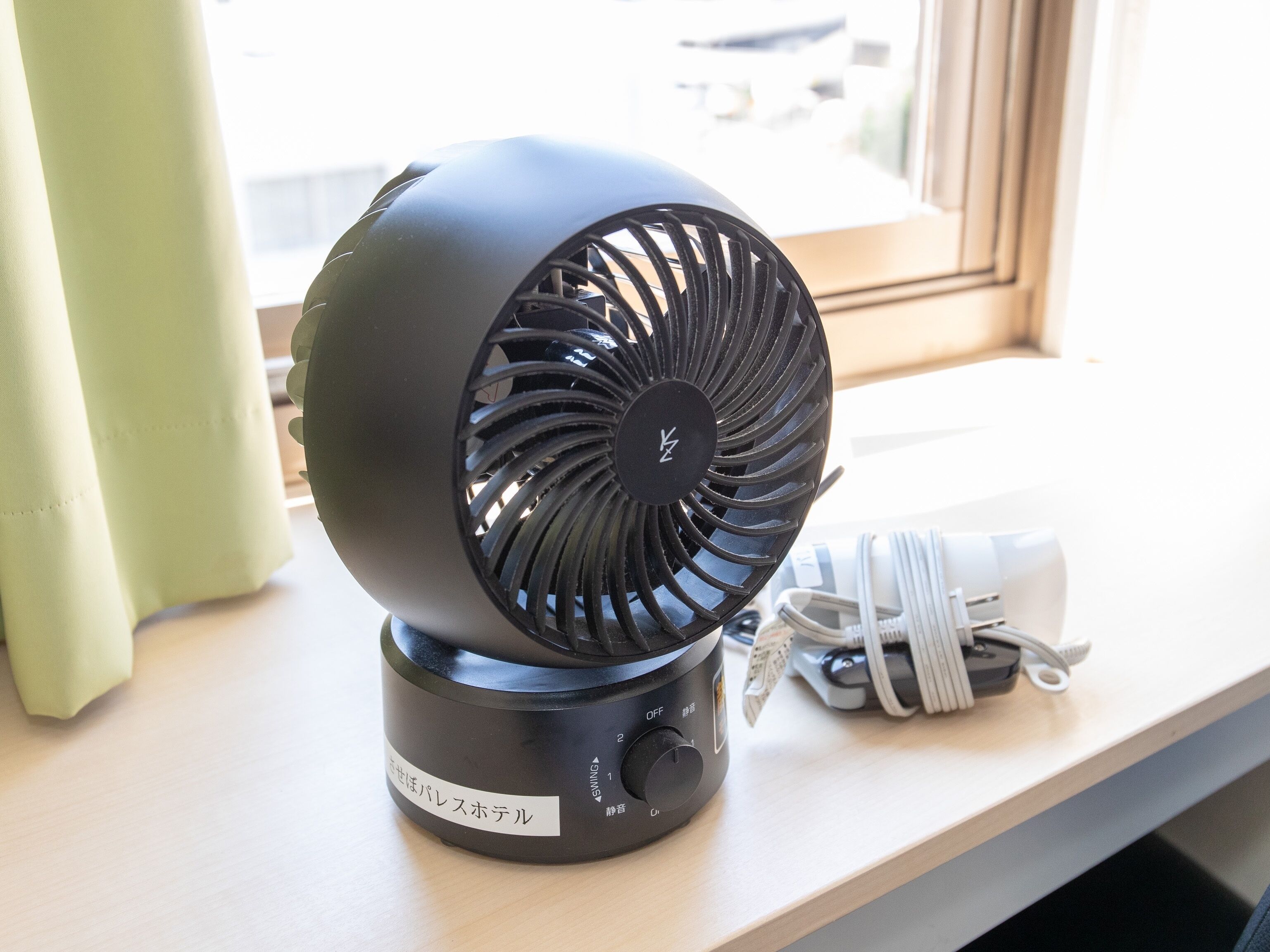A black portable fan with a label in a foreign language, placed on a wooden surface near a window.