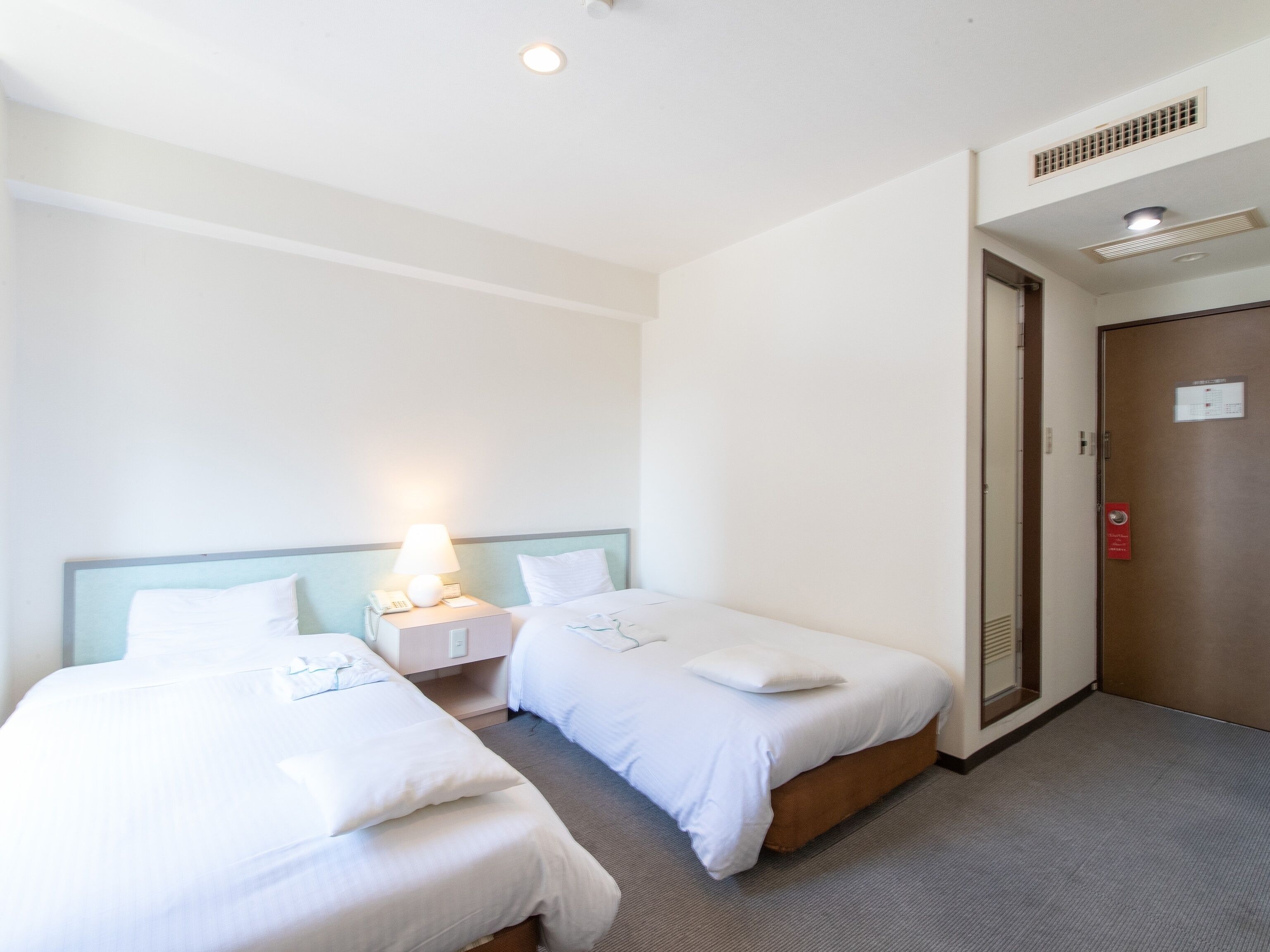 A hotel room with two single beds, a nightstand with a lamp, a door with a red emergency exit sign, and a ceiling vent.