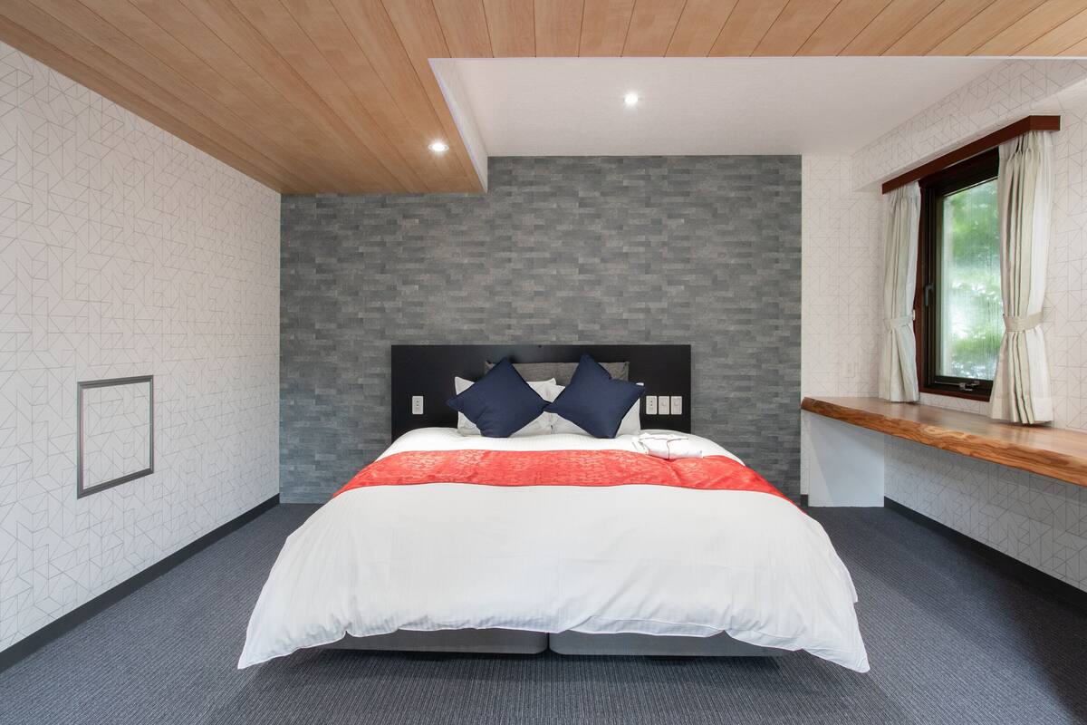 Une chambre moderne avec un plafond en bois, un mur en briques grises et un grand lit recouvert d’une couette blanche et rouge.