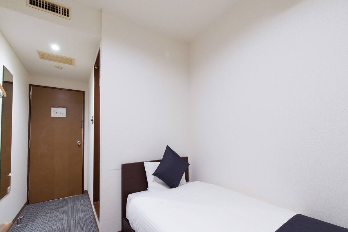 Une chambre d’hôtel avec un lit simple, une porte avec une pancarte, un sol recouvert de moquette et une bouche d’aération au plafond.