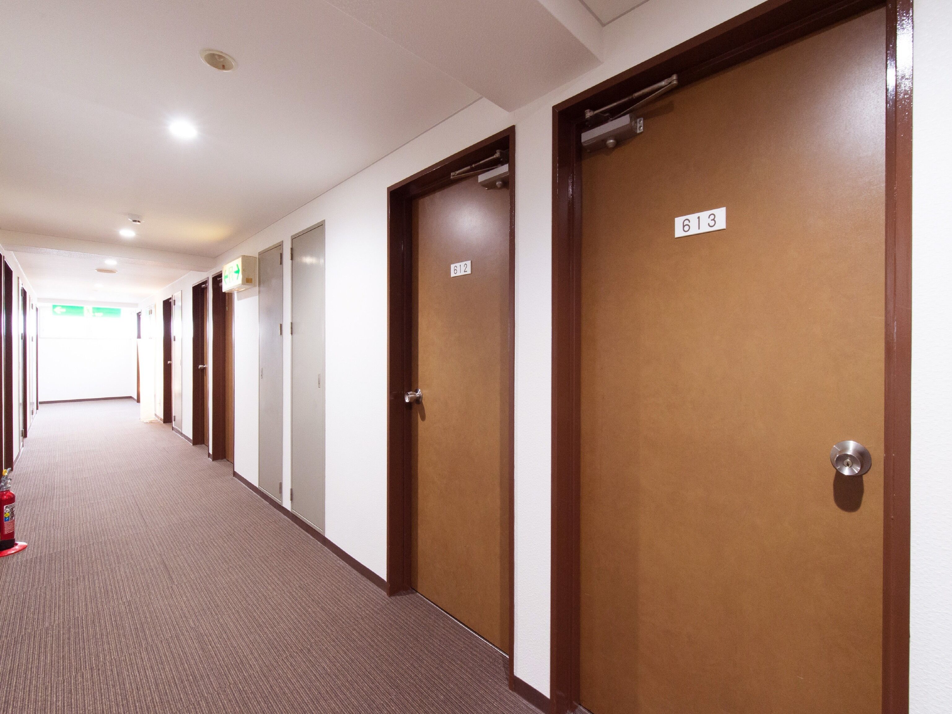 Un couloir d’hôtel avec les portes numérotées 612 et 613, un sol recouvert de moquette et un extincteur placé sur le côté gauche.