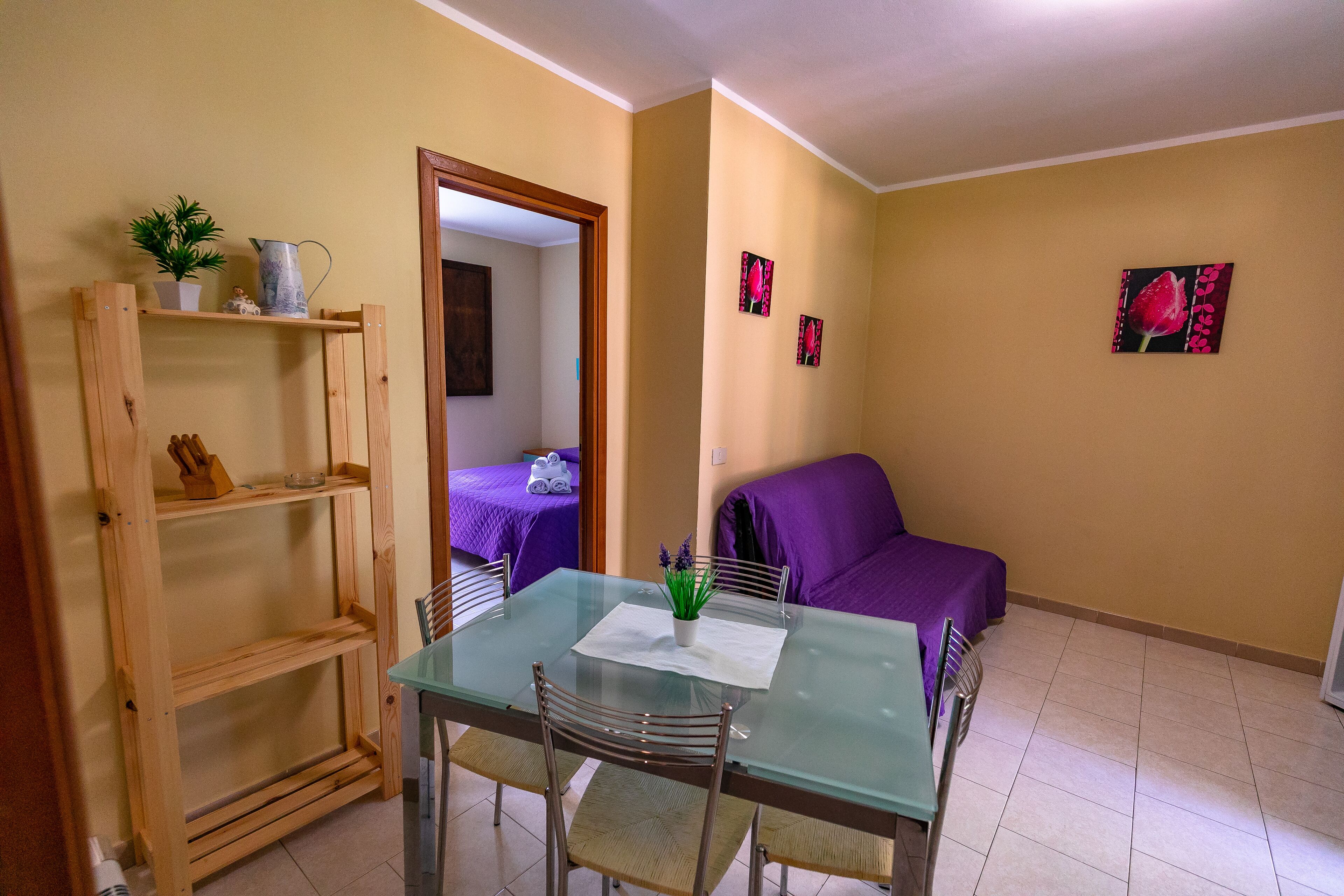 A small, cozy room with a glass dining table, two chairs, a wooden shelf unit, a purple sofa, and a bed with a purple cover.