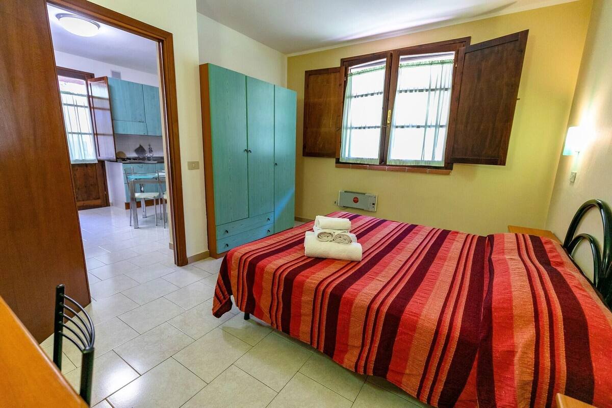 Un dormitorio con cama, ventana con contraventanas de madera, una kitchenette al fondo y una puerta que da a otra habitación.