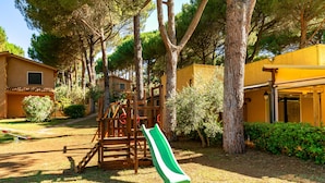 A playground with a slide and climbing structures, surrounded by trees and a yellow building. - Residence Il Paradiso (Guardistallo)