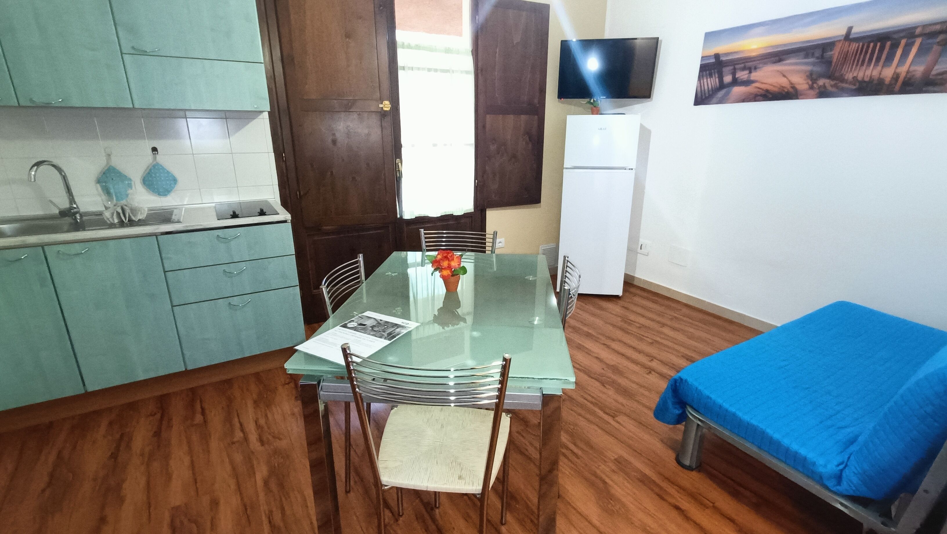 A compact kitchen with a dining table, chairs, a small refrigerator, and a TV on the wall.