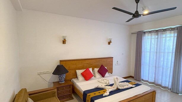 A hotel room with a wooden bed, a ceiling fan, a bedside table with a lamp, and a window with curtains. - Serendib Beach Resort (Induruwa)