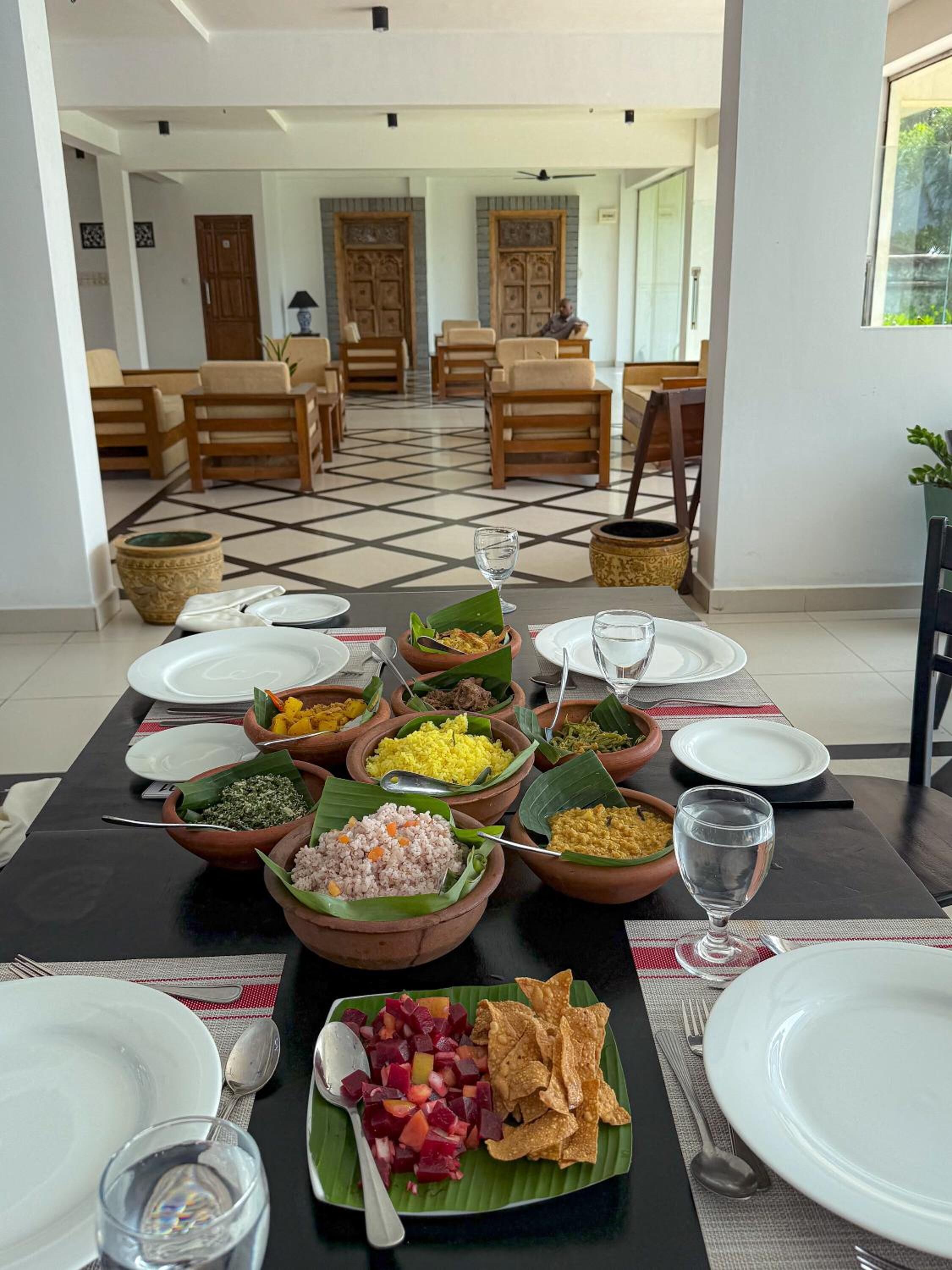 A dining table set with various dishes and drinks, surrounded by chairs and a view of a spacious room with a tiled floor and wooden furniture.