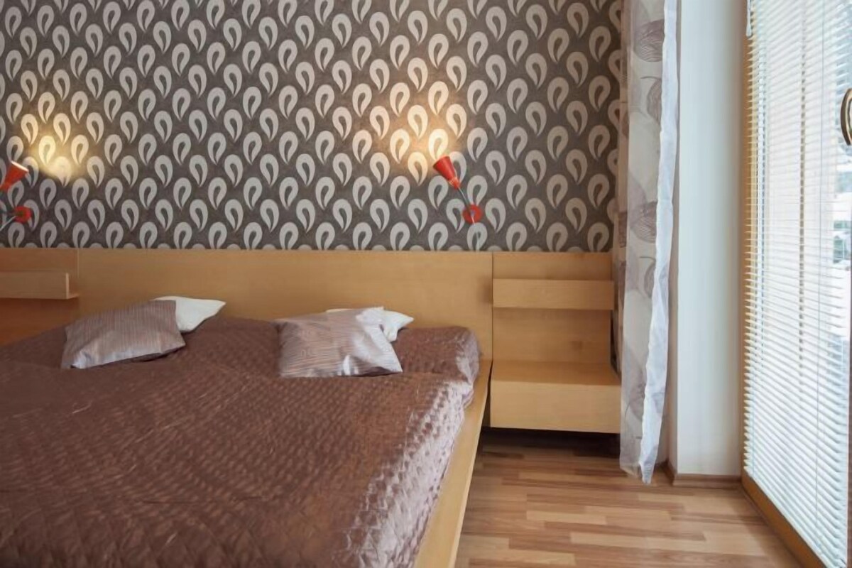 Une chambre à coucher avec un papier peint à motifs, une tête de lit en bois, un lit avec des oreillers et une fenêtre avec des stores.