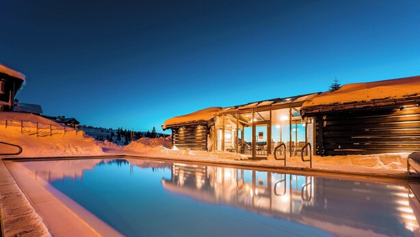 Una piscina al aire libre cubierta de nieve, con una cabaña de madera y un área de estar con paredes de cristal.