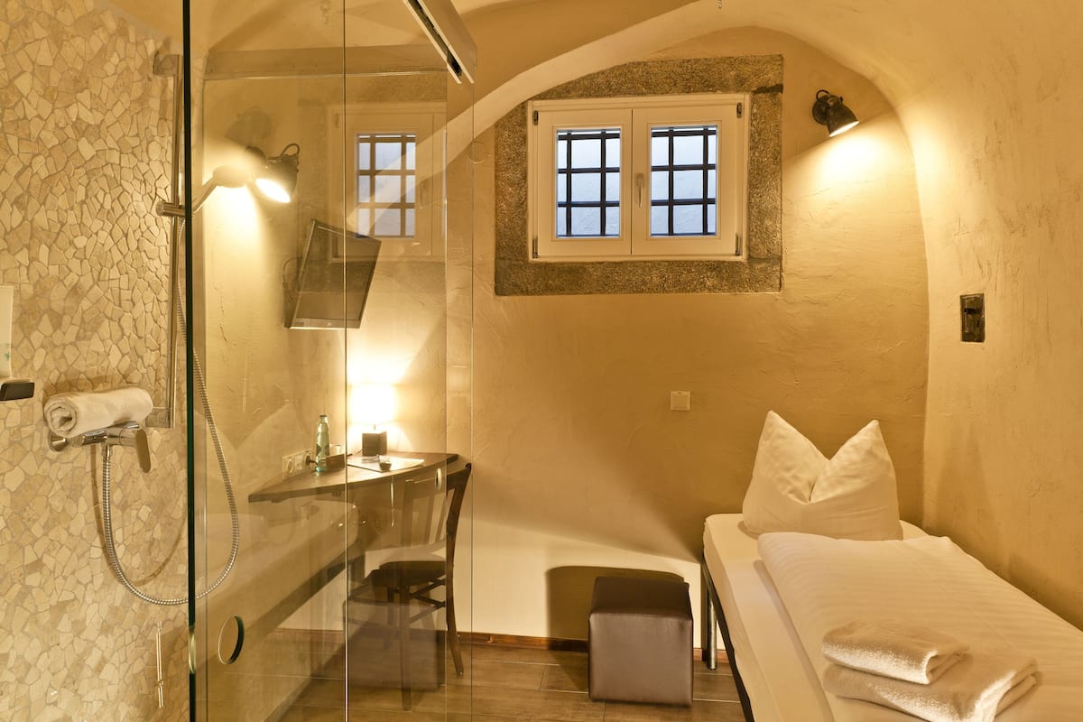 Una habitación pequeña con ducha, un escritorio, una cama y una ventana.