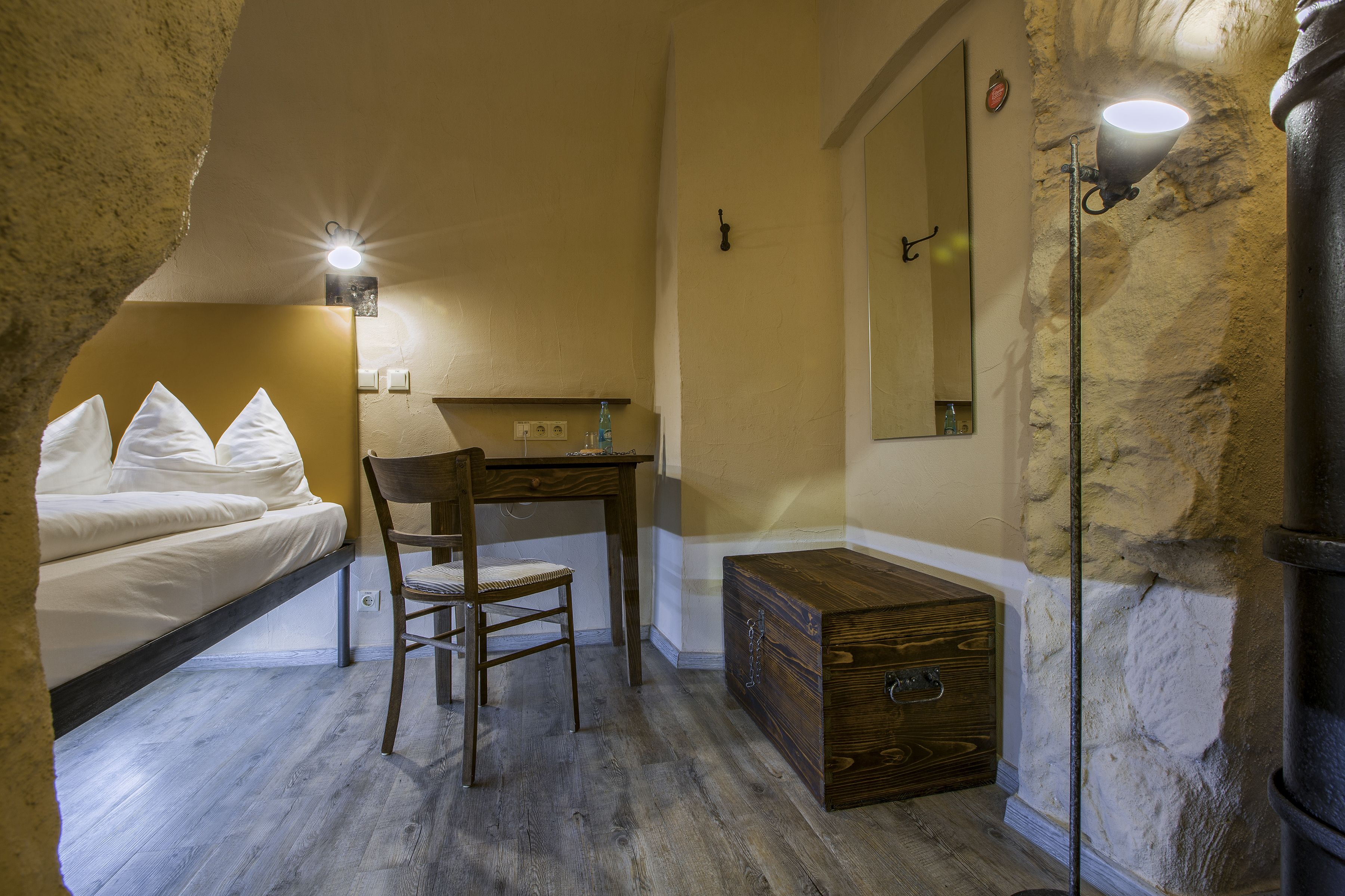 A bedroom with a bed, a desk, a chair, a wooden chest, and a nightstand with a lamp.