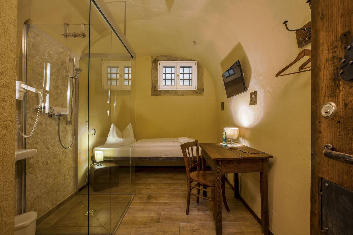 Habitación de hotel pequeña con ducha, cama, una mesita con silla, lámpara de noche y puerta de madera.