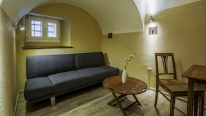 Una sala de estar acogedora con un sofá, una mesa de centro de madera y una silla. Se aprecia una ventana con vistas, una lámpara fijada a la pared y un pequeño marco de fotos.