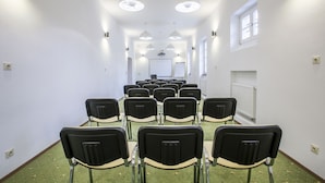 Un salón de conferencias con filas de sillas, paredes blancas e iluminación en el techo.