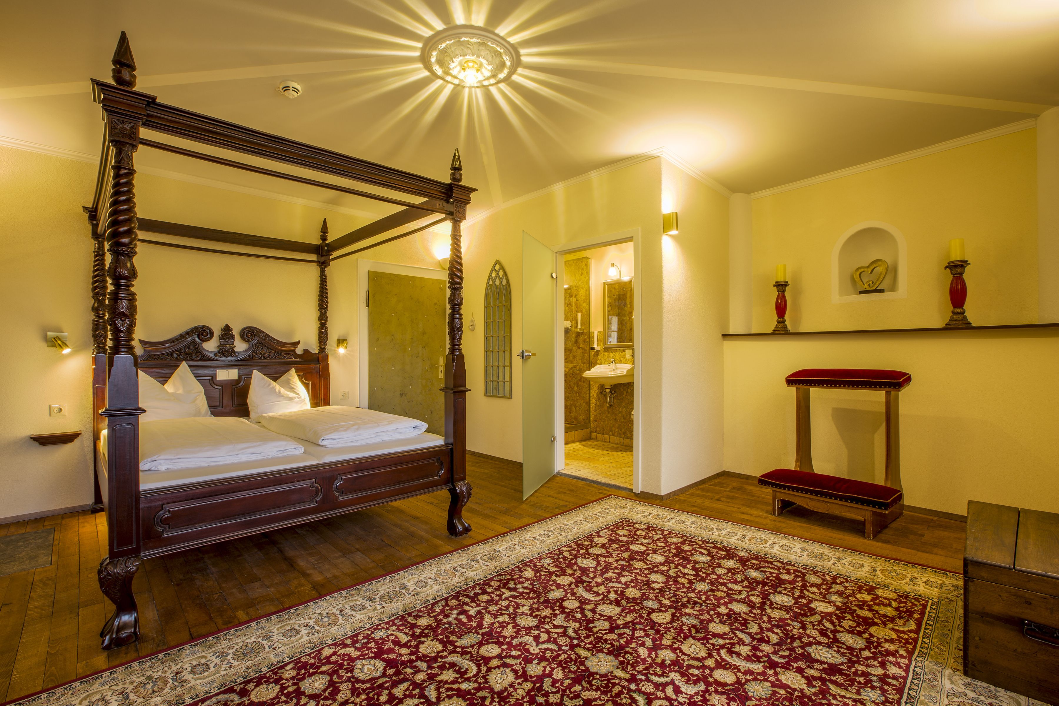 Una cama con dosel y cabecera de madera, una alfombra de diseño rojo y beige, un banco de madera y una pequeña mesa con un espejo.