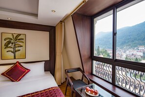 A hotel room with a bed, a desk with a plate of apples, a chair, and a large window offering a mountain view. - Freesia Hotel (Sa Pa)