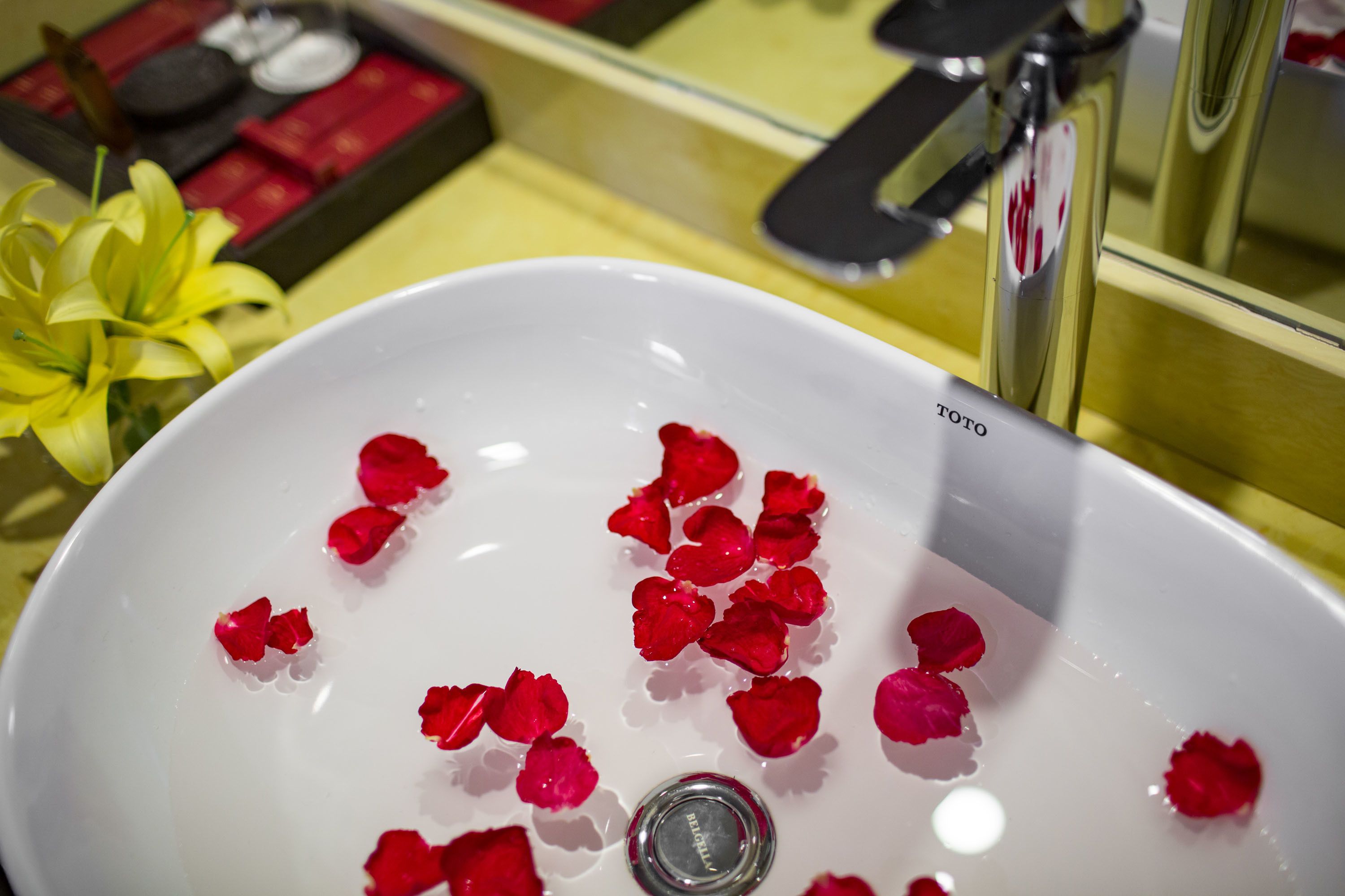 Un lavabo blanco con pétalos de rosa roja flotando en el agua, una flor amarilla y un espejo que refleja parte de la habitación.