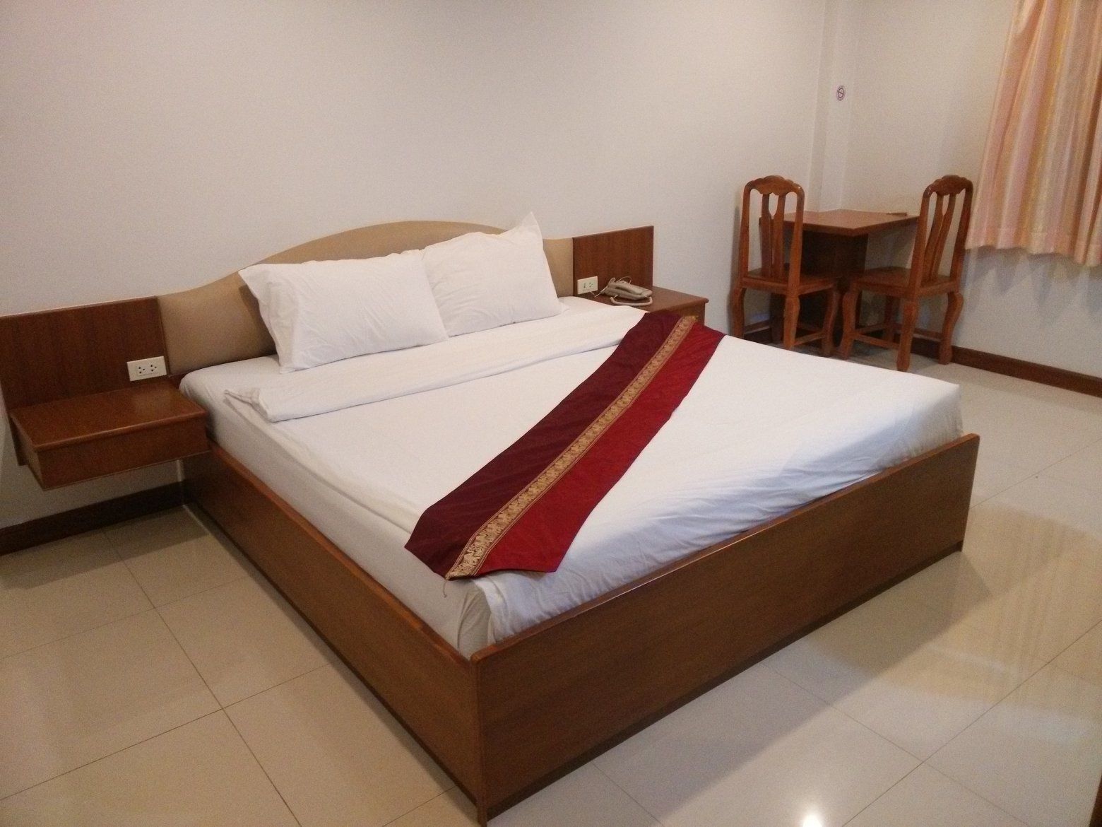 A neatly made bed with white linens and a red decorative strip, a wooden nightstand with a phone, and a wooden chair in a hotel room.
