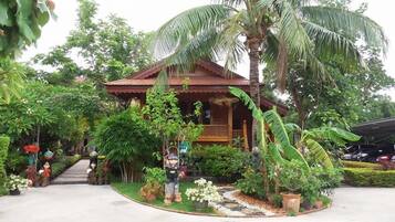 A traditional wooden house surrounded by lush greenery, including palm trees and various plants.