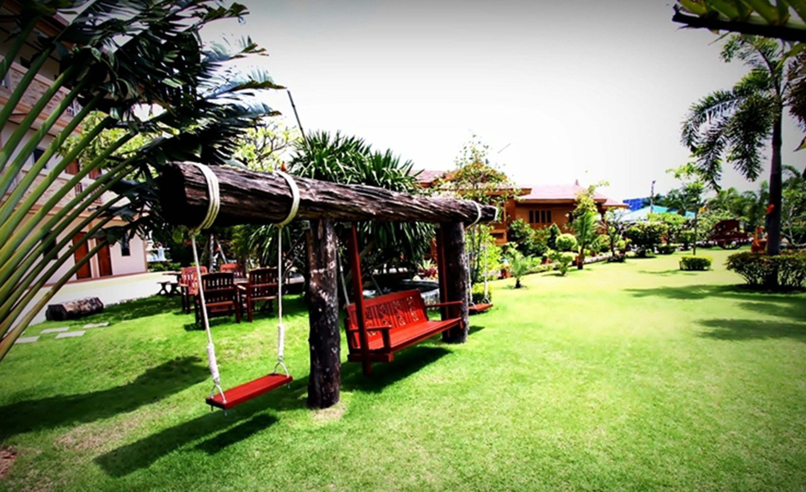 A wooden swing set in a well-maintained garden with a lawn, palm trees, and a building in the background.