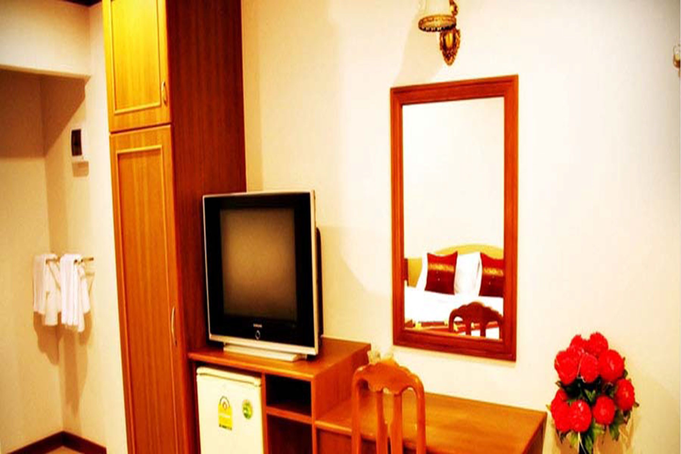 A hotel room with a television on a wooden stand, a small refrigerator, a wooden desk, a chair, a mirror on the wall, and a bouquet of red flowers.