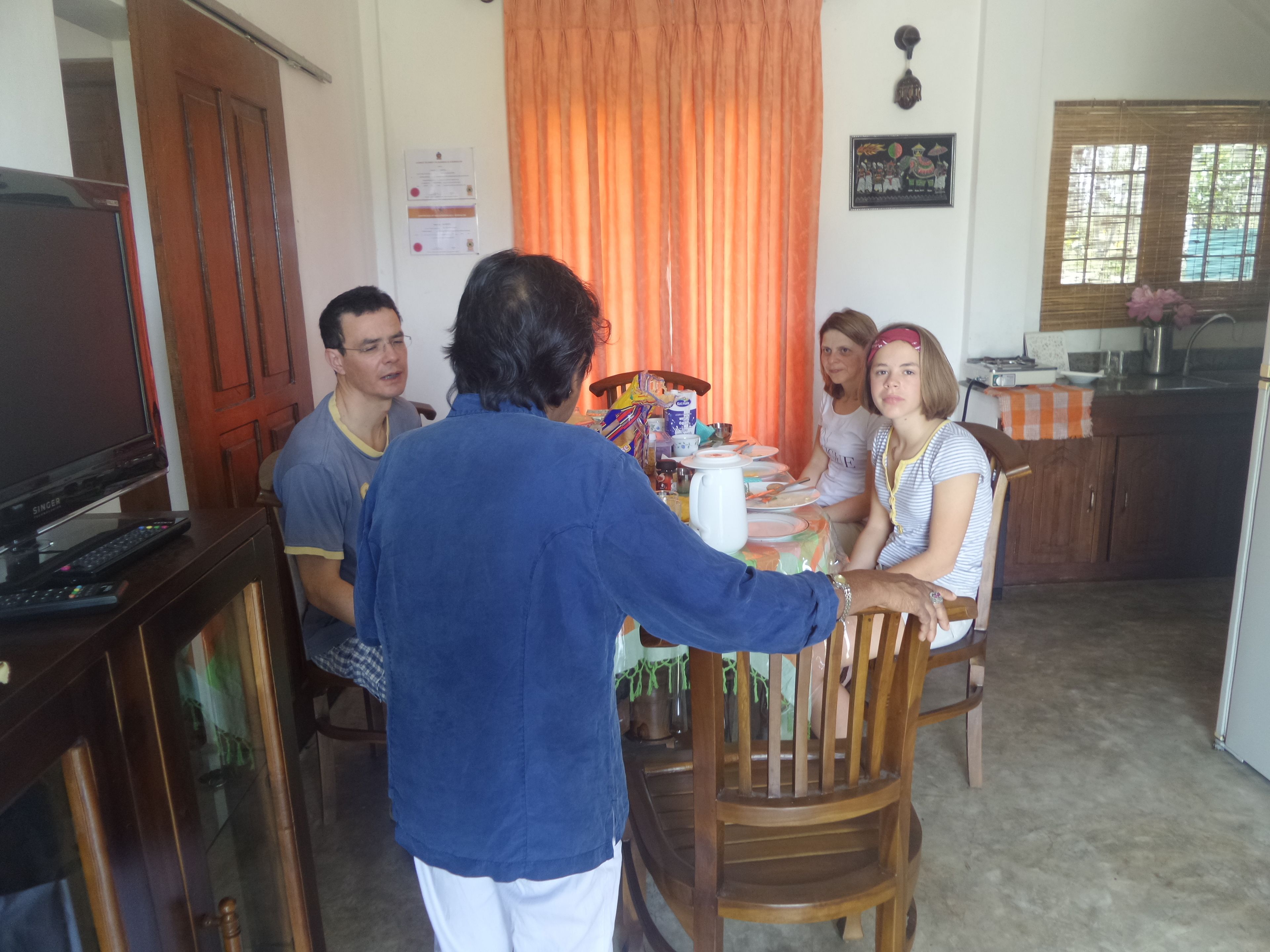 A family dining at a wooden table in a room with a kitchen and a TV.