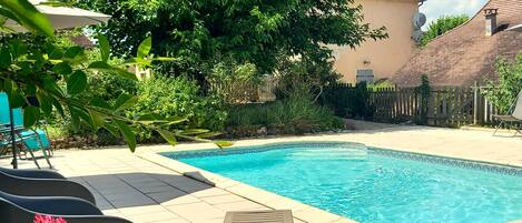 A swimming pool with a tiled deck, surrounded by greenery and a wooden fence.